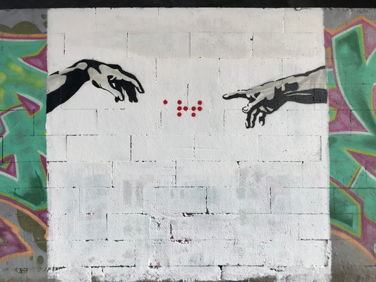 fresque de graffiti inspirée de La création d'Adam de Michel Ange, présentant les mains des personnages qui se tendent vers le mot art écrit en braille au centre de la fresque.