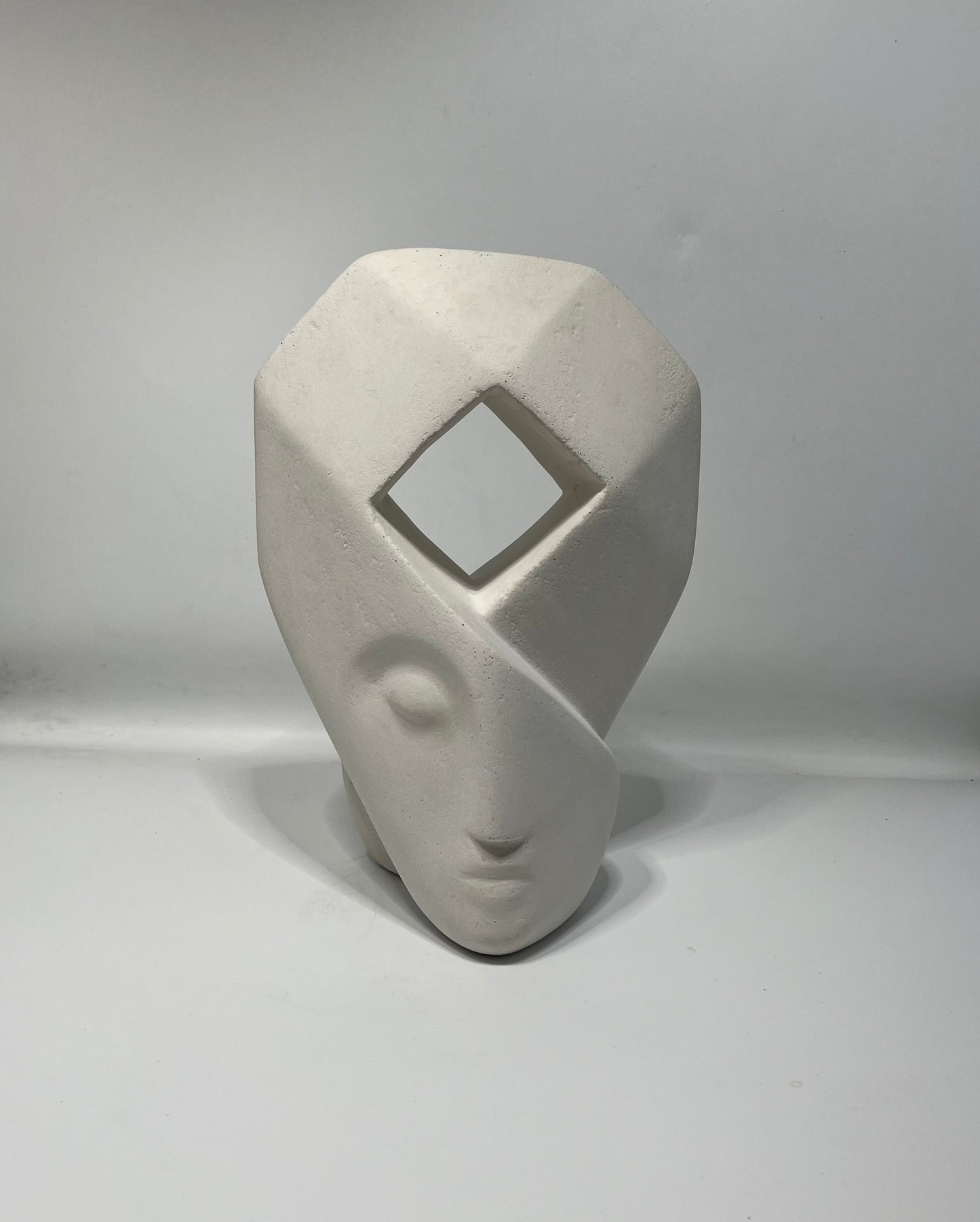 sculpture blanche d'un visage aux traits épurés dont le haut forme une coiffe géométrique avec une ouverture en son centre en forme de losange.