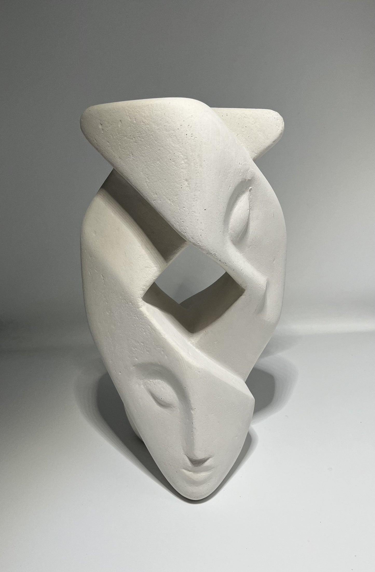 Sculpture blanche de deux visages imbriqués reposant l'un sur l'autre. Les visages sont stylisés et abstraits, avec des traits allongés. La sculpture est creuse, avec une ouverture carré en son centre.