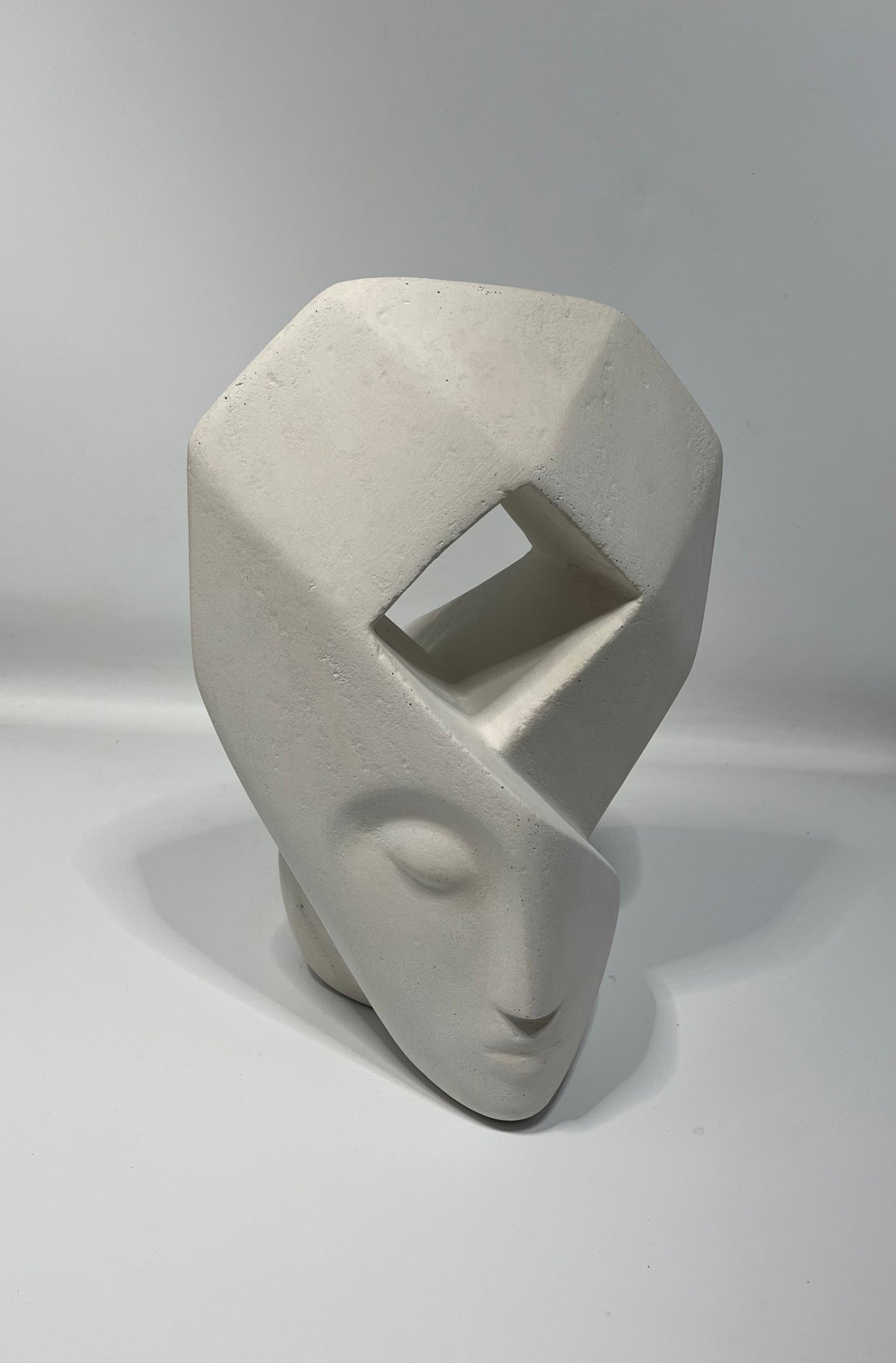 sculpture blanche d'un visage aux traits épurés dont le haut forme une coiffe géométrique avec une ouverture en son  centre en forme de losange. 